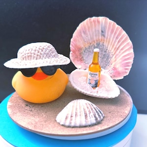 May include: A yellow rubber duck wearing sunglasses and a straw hat, with a miniature Corona Extra beer bottle in a seashell, set on a sandy beach scene with seashells. The scene is on a blue and white base.