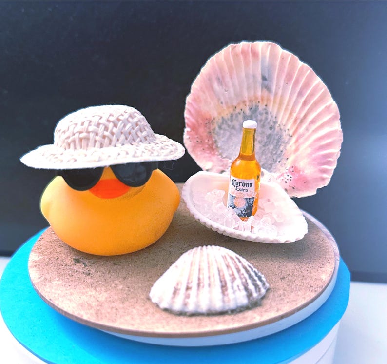 May include: A yellow rubber duck wearing sunglasses and a straw hat, with a miniature Corona Extra beer bottle in a seashell, set on a sandy beach scene with seashells. The scene is on a blue and white base.