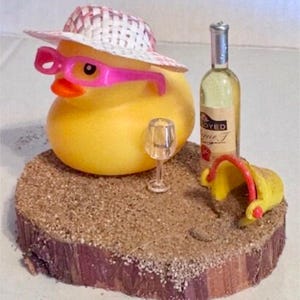 May include: A yellow rubber duck wearing a straw hat and pink sunglasses, with a wine bottle, a wine glass, and a yellow bucket on a wooden base covered in sand. The wine bottle has a label that says "OYED".