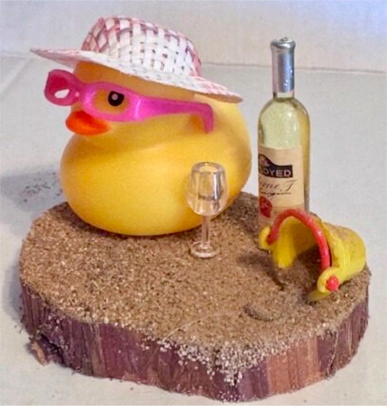 May include: A yellow rubber duck wearing a straw hat and pink sunglasses, with a wine bottle, a wine glass, and a yellow bucket on a wooden base covered in sand. The wine bottle has a label that says "OYED".