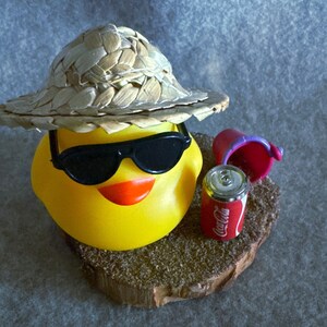 May include: A yellow rubber duck wearing black sunglasses and a straw hat, with an orange beak. Beside the duck is a small red Coca-Cola can and a pink bucket, all resting on a wooden base. The duck is a fun, summer-themed item.