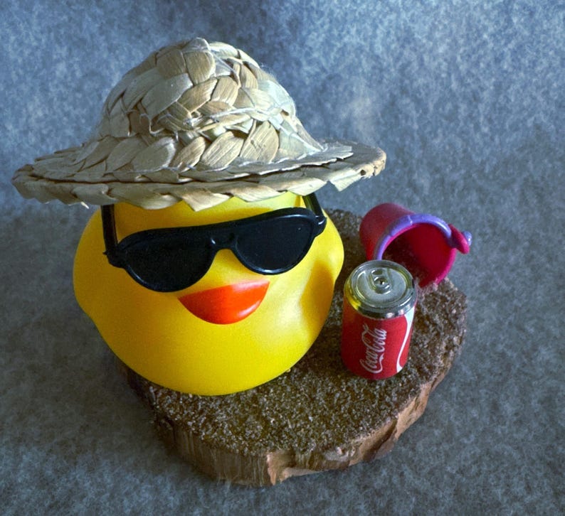 May include: A yellow rubber duck wearing black sunglasses and a straw hat, with an orange beak. Beside the duck is a small red Coca-Cola can and a pink bucket, all resting on a wooden base. The duck is a fun, summer-themed item.