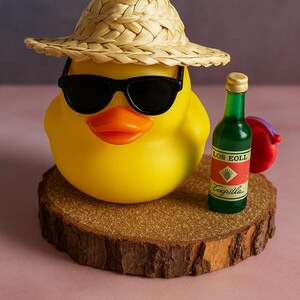 May include: A yellow rubber duck wearing black sunglasses and a straw hat, with an orange beak. Beside the duck is a small green bottle with a red label that reads "Los Eoll Cogilla". The duck is on a wooden slice.
