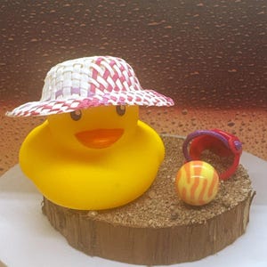 May include: A yellow rubber duck wearing a woven hat with pink and white accents. A small red and purple bucket and a striped ball sit on a sand-covered wooden base. The duck has an orange beak and is smiling.