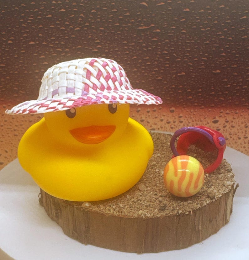 May include: A yellow rubber duck wearing a woven hat with pink and white accents. A small red and purple bucket and a striped ball sit on a sand-covered wooden base. The duck has an orange beak and is smiling.