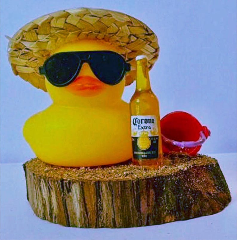 May include: A yellow rubber duck wearing sunglasses and a straw hat, with a bottle of Corona Extra beer and a small red bucket. The duck is resting on a wooden base, creating a fun, summery scene.