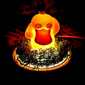 Può includere: Una lampada luminosa del personaggio Psyduck di colore giallo con un ampio becco arancione e grandi occhi, posizionata al centro di una base circolare ricoperta di muschio verde scuro. La luce emette un bagliore giallo caldo su sfondo scuro.