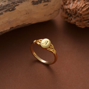 May include: A gold ring with an oval-shaped center piece featuring the letter "A" engraved inside. The ring has a delicate, intricate design.
