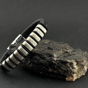 May include: A black leather bracelet with silver-colored rectangular accents. Each accent features engraved text. The bracelet has a braided design and a silver clasp. The bracelet is displayed on a dark, textured surface.