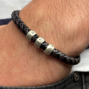 May include: Braided dark brown leather bracelet with three silver beads. The beads are engraved with the names "Eva", "Johe", and the number "0257". The bracelet is worn on a wrist.