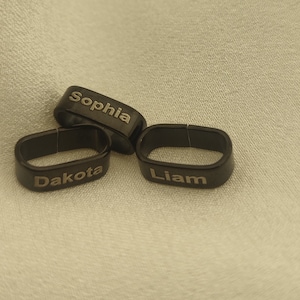 May include: Three black personalized rings with the names Dakota, Sophia, and Liam engraved in gold lettering.