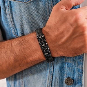 May include: A black braided leather bracelet featuring five rectangular black metal beads. Each bead is engraved with white Arabic text. The bracelet has a secure clasp mechanism and is worn around the wrist, showcasing a textured design.