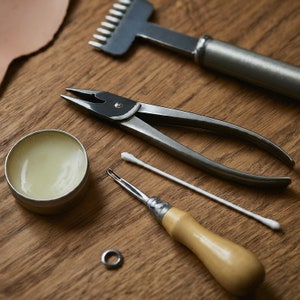 May include: A set of leatherworking tools on a wooden surface. The tools include a leather punch, a pair of pliers, a stitching awl, a cotton swab, and a small container of leather conditioner.