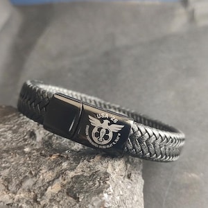Könnte beinhalten: Schwarzes geflochtenes Lederarmband mit rechteckigem Verschluss. Der Verschluss ist mit "USA '26" und einem Fußball-Emblem graviert. Das Armband liegt auf einer grauen Steinoberfläche.