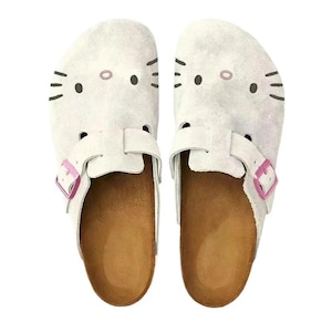 Könnte beinhalten: Ein Paar weiße Clogs mit einem Cartoon-Katzen-Gesicht-Design. Die Clogs haben ein hellbraunes Fußbett, eine rosa Schnalle und schwarze Details. Die Schuhe sind im Slip-On-Stil gestaltet.