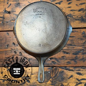 May include: A vintage cast iron skillet with a long handle and a circular cooking surface. The skillet is marked "Wagner Ware Sidney -0- 9A" and is set against a wooden background. The skillet is a dark, metallic color.