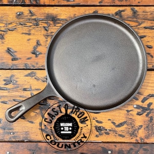 May include: A black cast iron griddle pan with a long handle and a round cooking surface. The pan is sitting on a wooden surface. The handle has a hole for hanging. The logo "CAST IRON COUNTRY" is visible.