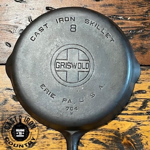 May include: A cast iron skillet with the text "CAST IRON SKILLET 8 GRISWOLD ERIE PA U.S.A." The skillet is dark gray and has a handle. The skillet is on a wooden surface.