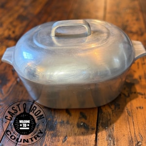 May include: A vintage aluminum cooking pot with a lid and handle. The pot has side handles and a rounded shape. The lid has a single handle. The pot is sitting on a wooden surface. The text "CAST IRON COUNTRY" is visible.