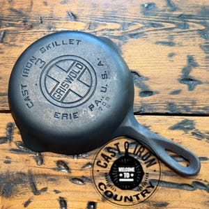 May include: A black cast iron skillet with a handle, featuring the text "GRISWOLD ERIE PA, U.S.A." and "CAST IRON SKILLET". The skillet is on a wooden surface. A circular logo with the text "CAST IRON COUNTRY" is in the bottom right corner.