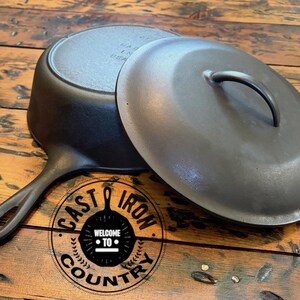 May include: A black cast iron skillet with a lid, resting on a wooden surface. The skillet has a long handle and a circular base. The lid has a curved handle. The image includes a logo that says "Cast Iron Country".