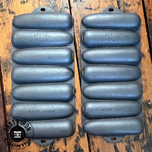 May include: Two cast iron Wagner Ware loaf pans. Each pan has a series of rounded loaf molds and a small hole at the top for hanging. The text "WAGNER WARE" and "MADE IN USA" are visible on the pans.