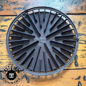 May include: A round, dark gray cast iron trivet featuring a radial spoke design with raised ridges extending from the center. The center is embossed with the text PAT'D JUNE 27 1911 and the number 13. It has a circular outer rim and small feet.
