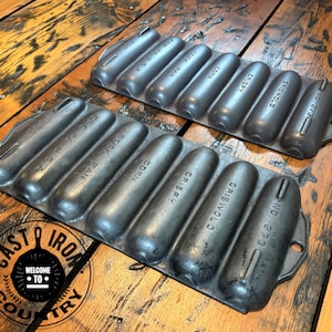 May include: Two dark gray cast iron corn stick pans with seven molded cavities each. The pans feature embossed text reading GRISWOLD, CRISPY, CORN, STICK PAN, ERIE PA U.S.A., and NO. 273. The metal surfaces have a matte finish with visible texture.