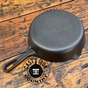 May include: A black cast iron skillet with a long handle and a circular cooking surface. The skillet has the text "Griswold Erie PA" and "No. 6" embossed on the bottom. The image also includes a logo that says "Cast Iron Country".