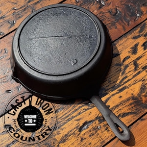 May include: A black cast iron skillet with a long handle and a circular cooking surface. The skillet is on a wooden surface. The text "CAST IRON COUNTRY" is in a circular logo.