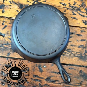 May include: A well-seasoned, black cast iron skillet with a single handle. The number "9" is embossed on the cooking surface. The skillet is on a wooden surface. The logo "Cast Iron Country" is visible.