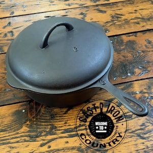 May include: A black cast iron skillet with a lid, handle, and a circular logo that says "Cast Iron Country". The skillet is on a wooden surface.