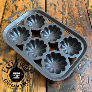 May include: A cast iron pan with six shell-shaped molds. The pan has a rectangular shape with handles on each side. The pan is dark gray and sits on a wooden surface. The text "CAST IRON COUNTRY" is visible.