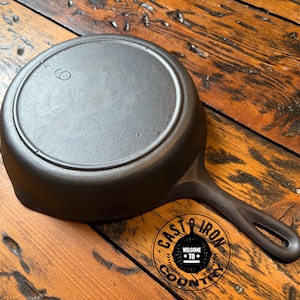 May include: A dark gray cast iron skillet shown from the underside, featuring a circular heat ring and the number 6 embossed on the base. The handle includes a teardrop-shaped hole at the end for hanging. The surface has a matte finish.