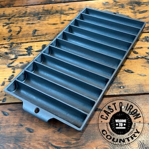 May include: A rectangular black cast iron corn stick pan featuring seven long, narrow, semi-cylindrical molds arranged in parallel rows. The pan includes two small circular holes at opposite ends for hanging and a textured, dark metallic finish.
