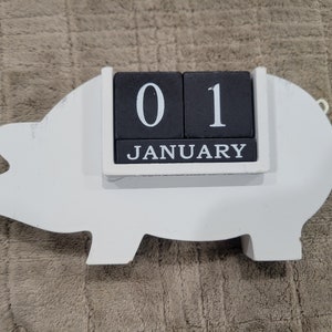 May include: White wooden pig figurine with a black and white calendar block that reads "01 JANUARY".
