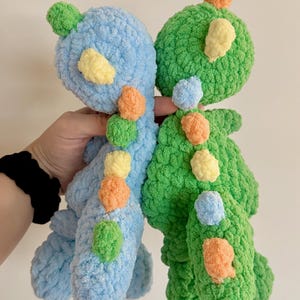 May include: Two crocheted dinosaur plushies, one blue and one green, with colorful accents.