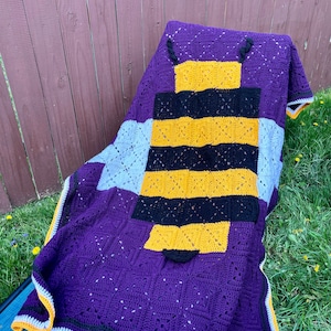 May include: A crocheted blanket with a bee design. The bee is made with yellow, black, and white yarn. The blanket is purple, white, and yellow.