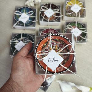 May include: A collection of small, round ceramic bowls featuring intricate, colorful floral patterns. Each bowl is individually packaged in a clear square box, tied with white twine, and includes a white diamond-shaped tag with a printed name.
