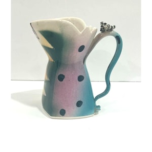 May include: A ceramic pitcher featuring a teal and purple gradient body with black polka dots. The pitcher has a flared, jagged rim and a curved handle topped with a small black and white chequered accent piece. The design includes triangular shapes.