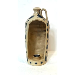 Może przedstawiać: Beżowy dzbanek ceramiczny z dużym pionowym wycięciem z przodu, wyposażony w uchwyt u góry oraz ciemnoniebieskie malowane wzory w liście i kropki. Wnętrze dzbanka posiada poziome żłobienia oraz okrągły otwór w podstawie na świecę.
