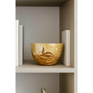 Op de afbeelding: Een decoratieve keramische kom met een landschapsontwerp in bruin- en beigetinten. De kom heeft een boom, glooiende heuvels en een zon. De kom staat op een plank tussen twee witte boeken.