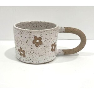 Könnte beinhalten: Ein Keramikbecher mit gesprenkelter weißer Glasur und braunen, handgemalten Blumenmotiven. Der Becher hat einen zylindrischen Körper und einen dicken, geschwungenen braunen Henkel. Die Außenfläche wirkt texturiert mit kleinen braunen Sprenkeln.