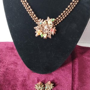 May include: A vintage gold-toned necklace and matching earrings set. The necklace features a floral pendant with colorful enamel flowers and a triple-strand beaded chain. The earrings mirror the pendant design. The jewelry is displayed on a black velvet background.