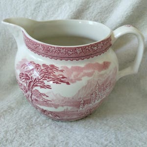May include: A cream-colored ceramic creamer pitcher featuring a detailed transferware pattern in a deep pink hue. The design depicts a landscape with a large tree, a castle, and mountains. A decorative floral border encircles the top rim.