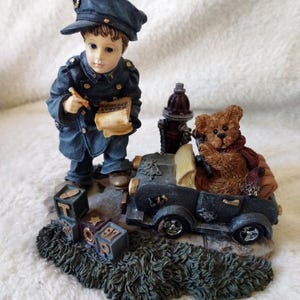 May include: A figurine featuring a child dressed in a dark blue uniform and cap, writing on a paper, standing next to a dark blue toy car with a brown teddy bear inside. A small red fire hydrant and three gray alphabet blocks are on the base.