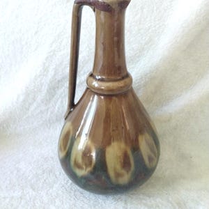 May include: A brown ceramic pitcher with a tall, slender neck and a single handle. The body features a rounded base decorated with a repeating pattern of cream and dark brown circular spots, finished with a glossy, reflective glaze.