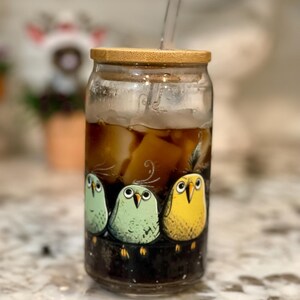 Puede incluir: Un tarro de cristal con tapa de bambú y una pajita de plástico transparente. El tarro está lleno de café helado y tiene un diseño de dibujos animados de colores con tres pájaros en la parte delantera.