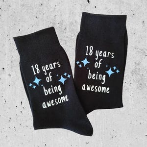 Op de afbeelding: Een paar zwarte sokken met de witte tekst "18 jaar geweldig zijn" en blauwe sterren. Deze sokken zijn gemaakt van zacht materiaal, perfect voor dagelijks gebruik.