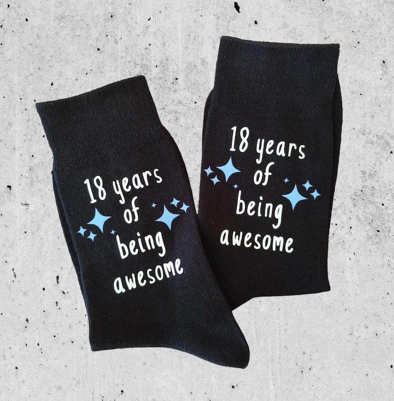 Op de afbeelding: Een paar zwarte sokken met de witte tekst "18 jaar geweldig zijn" en blauwe sterren. Deze sokken zijn gemaakt van zacht materiaal, perfect voor dagelijks gebruik.
